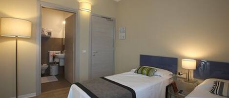 A hotel room with two beds, a bedside table with a lamp, a wardrobe, and a bathroom visible through an open door.