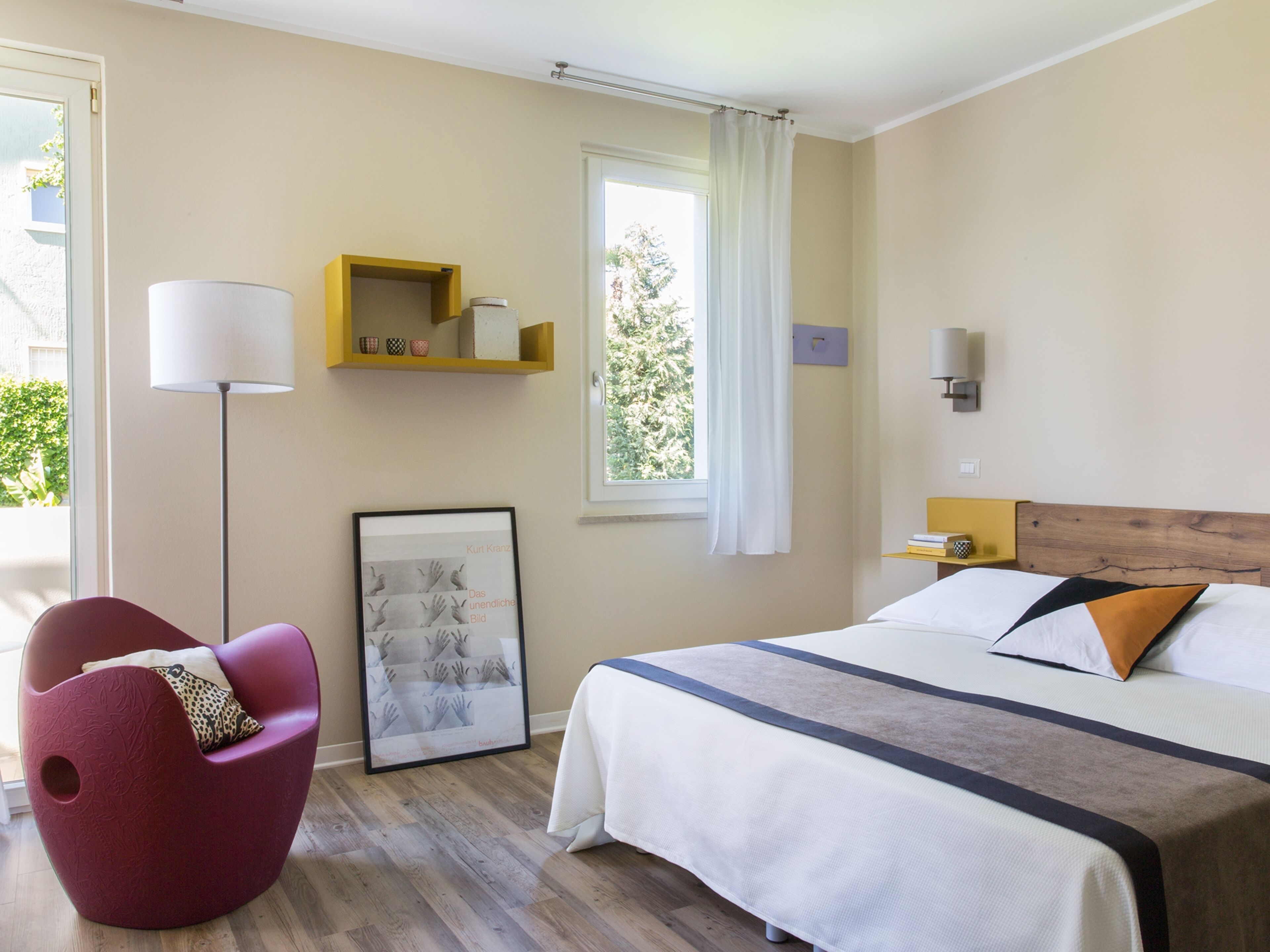 A hotel room with a bed, a chair, a bedside table, a lamp, a window, and a framed picture on the wall.