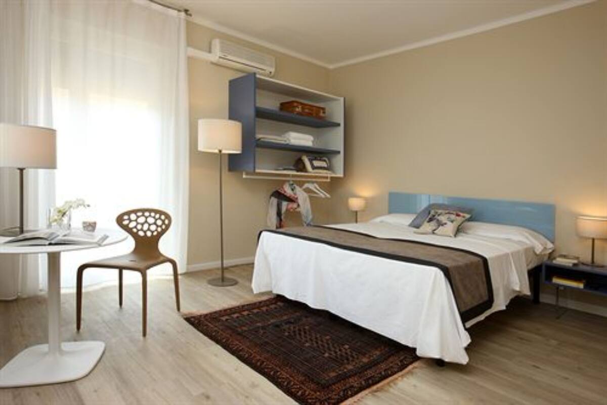 Una habitación de hotel con cama, mesita de noche, lámpara, silla y una estantería con objetos personales.