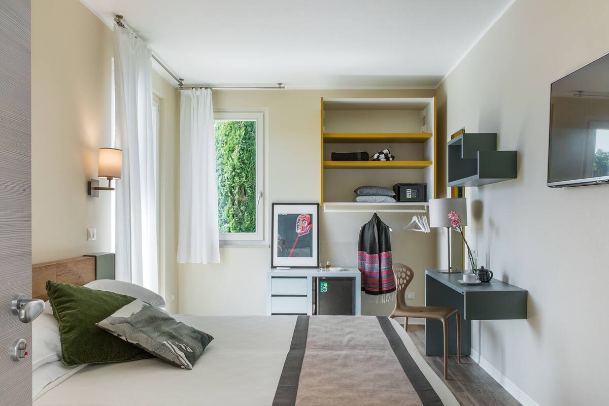 Habitación de hotel con cama, escritorio, silla, televisión y ventana con cortinas.