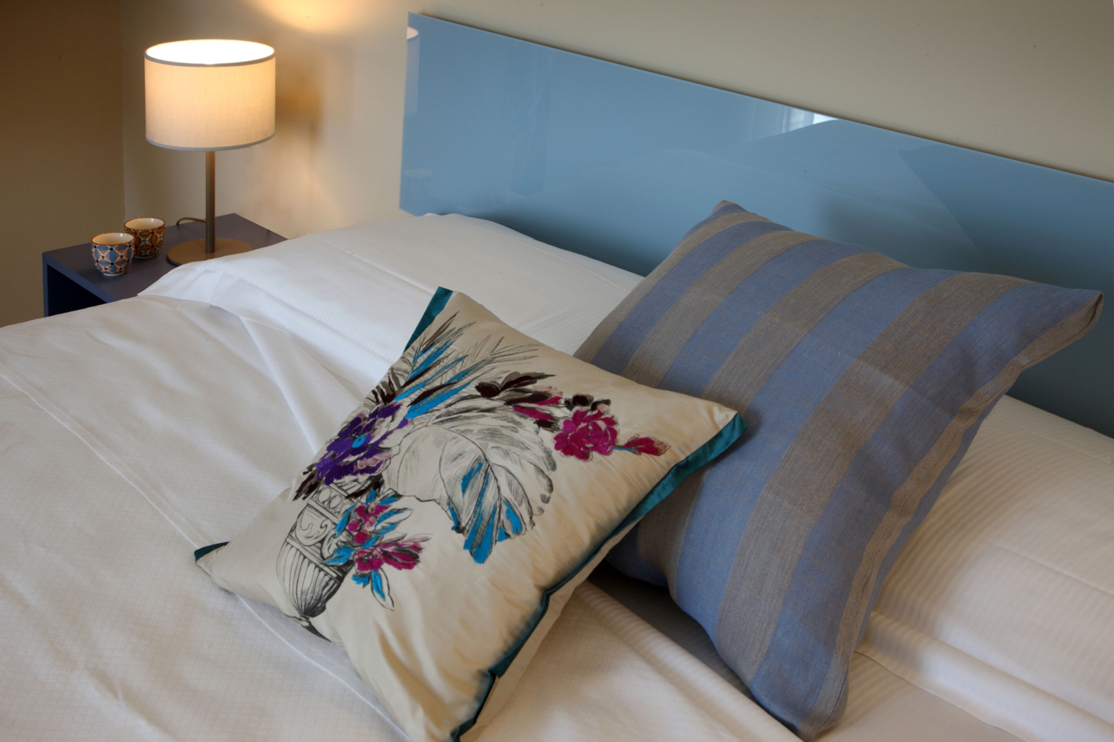 A neatly made bed with a floral pillow and striped pillow, a bedside lamp, and a small table with two cups.