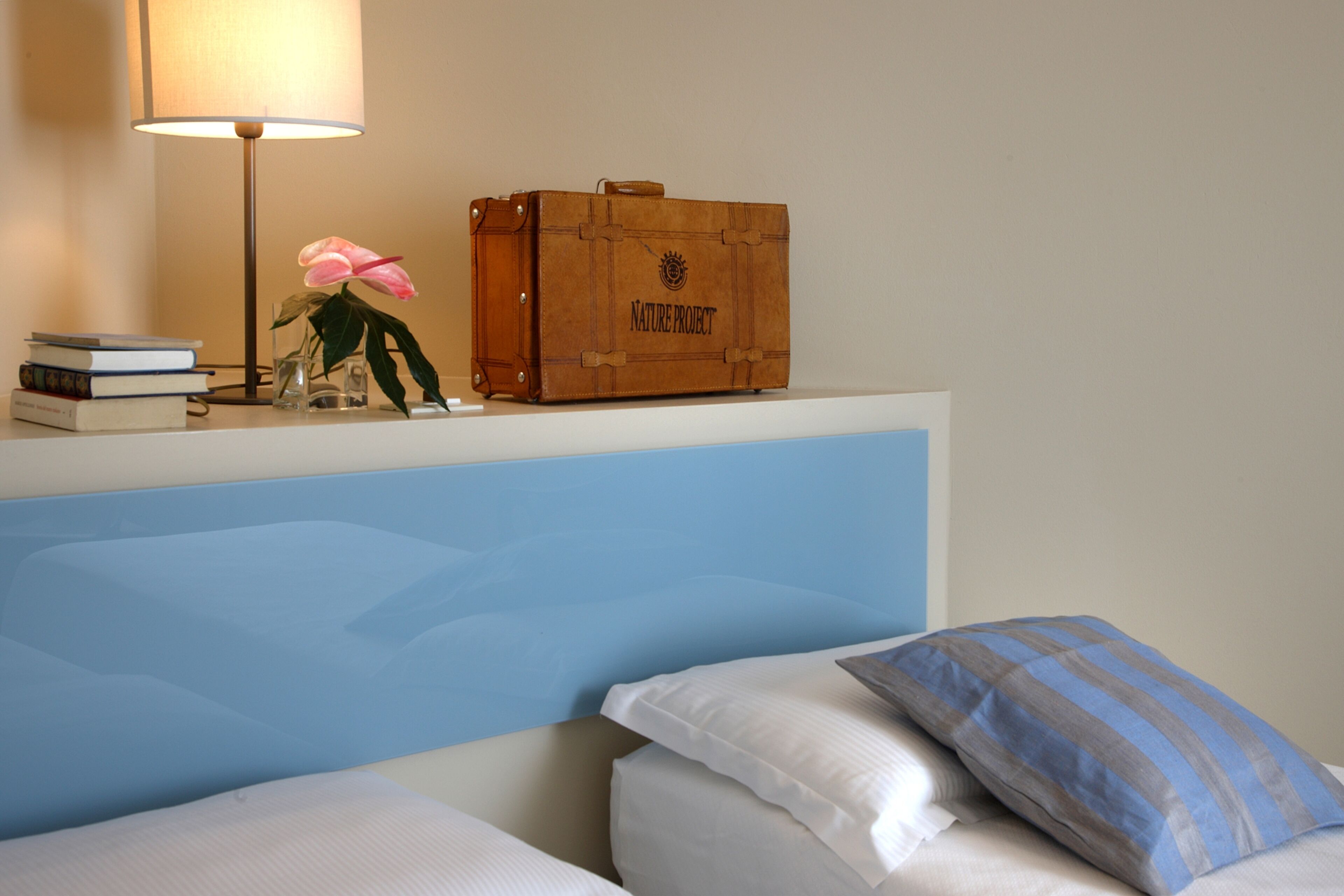 Un dormitorio con cama, almohadas, una mesita de noche con lámpara, libros y una maleta.