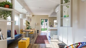 A modern living room with a wooden floor, a yellow ottoman, a colorful chair, and a view of the outdoors.