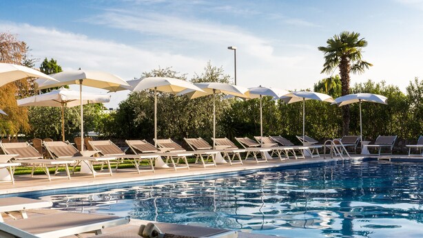 A poolside area with sun loungers and umbrellas, surrounded by greenery and a clear sky.