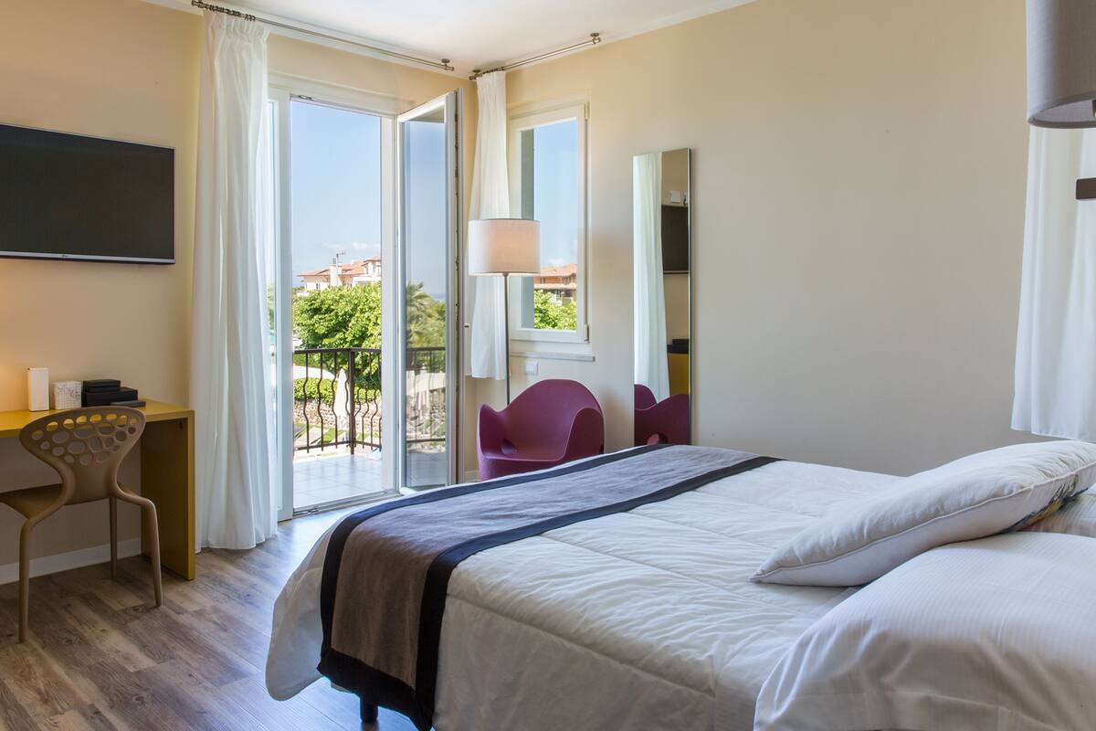 Habitación de hotel con cama, escritorio con silla, televisión y balcón con vistas a edificios y áreas verdes.