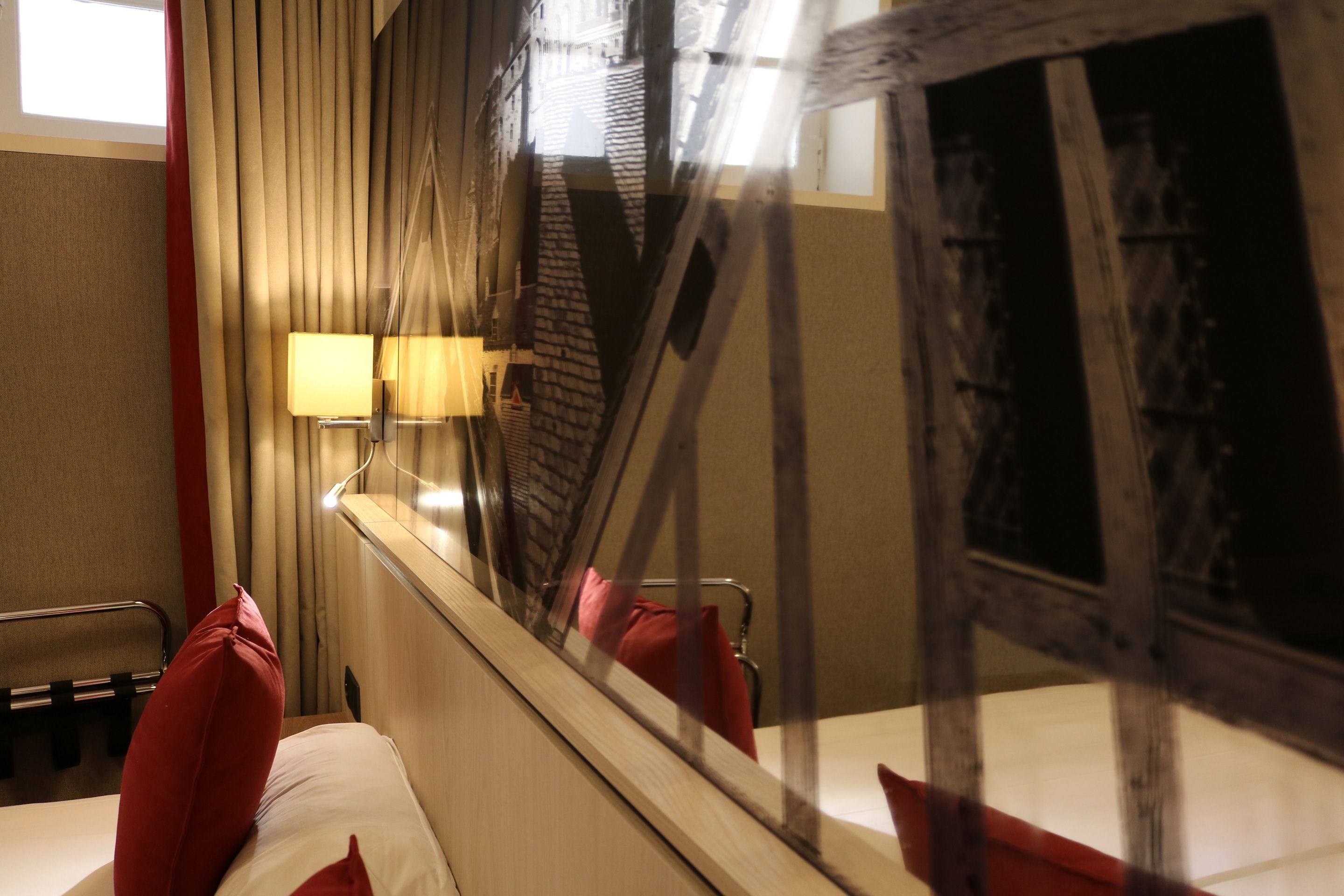 A hotel room with a bed, a window with curtains, a lamp, and a view of a building outside.