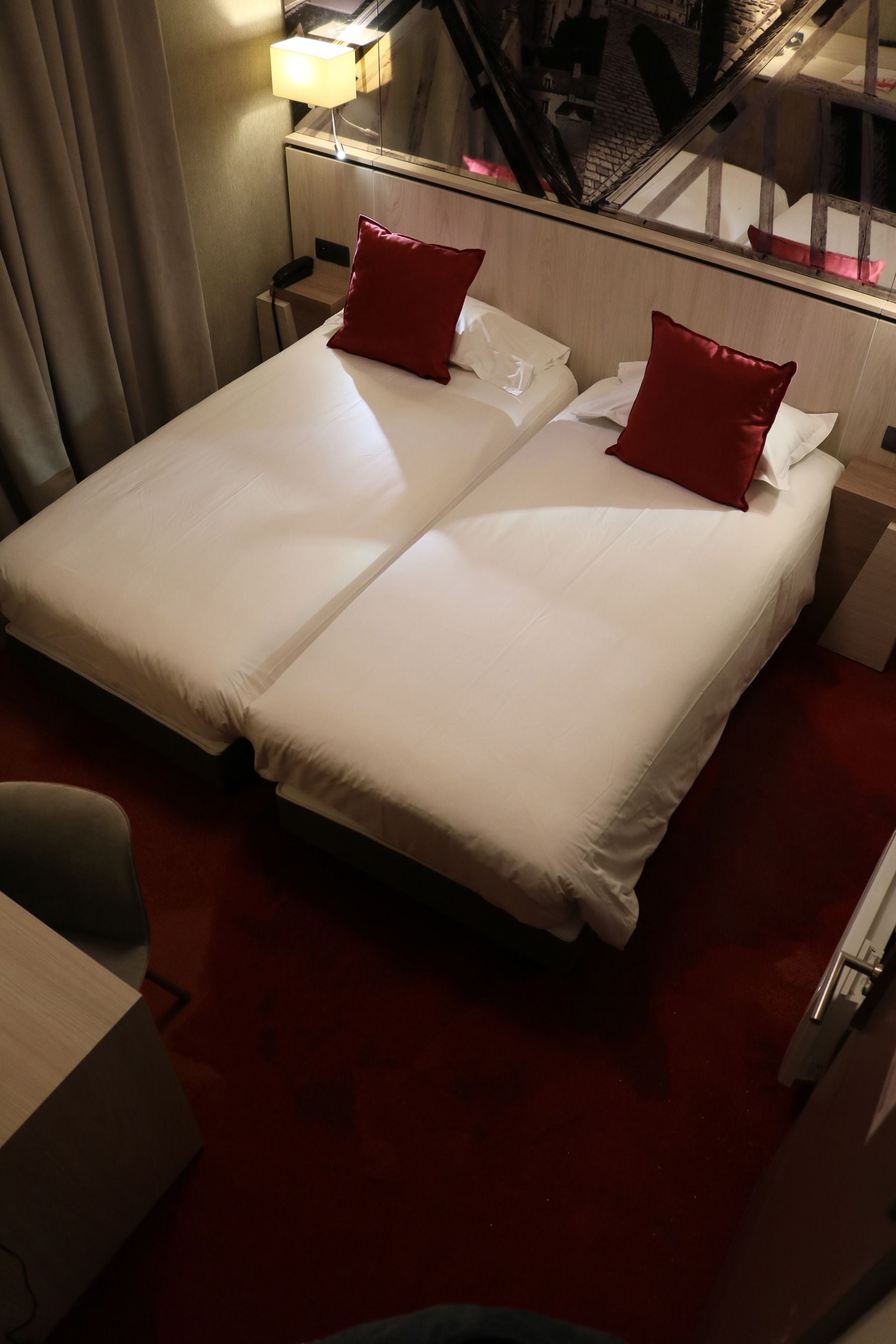 A hotel room with a double bed, two red pillows, a bedside table with a lamp, and a view of a cityscape through a window.