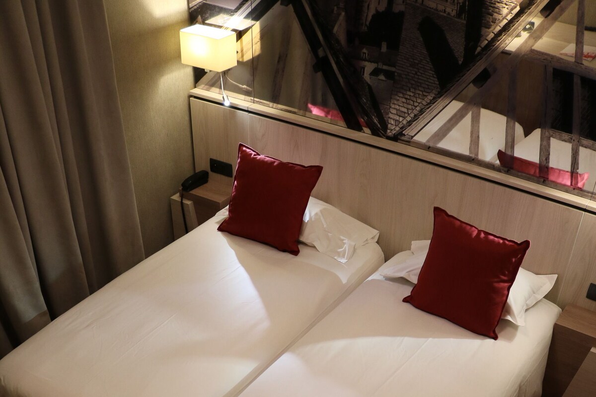 Habitación de hotel con cama doble, dos almohadas rojas, una mesita de noche, una lámpara y vistas a una escalera.