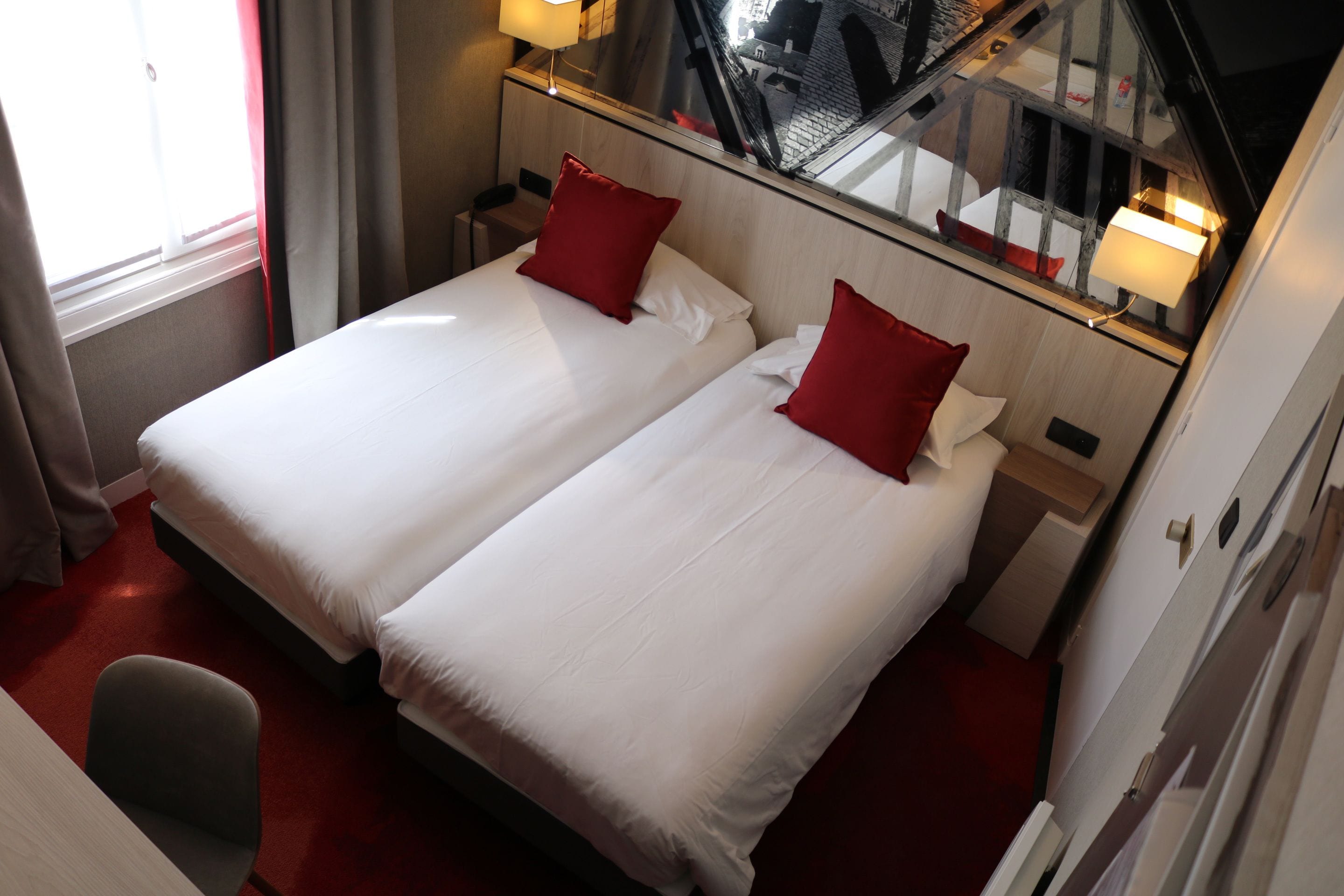 A hotel room with two beds, red pillows, a small desk, and a chair.