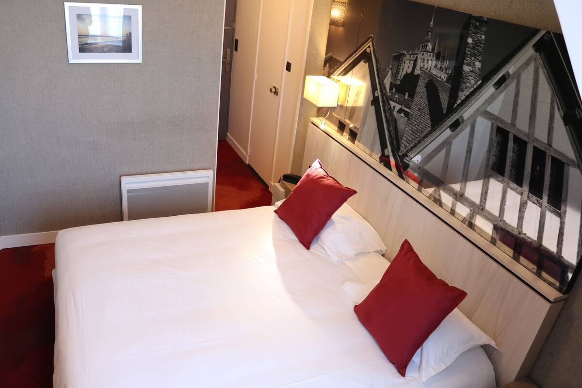 A hotel room with a bed, red pillows, a nightstand with a lamp, and a cityscape mirror.