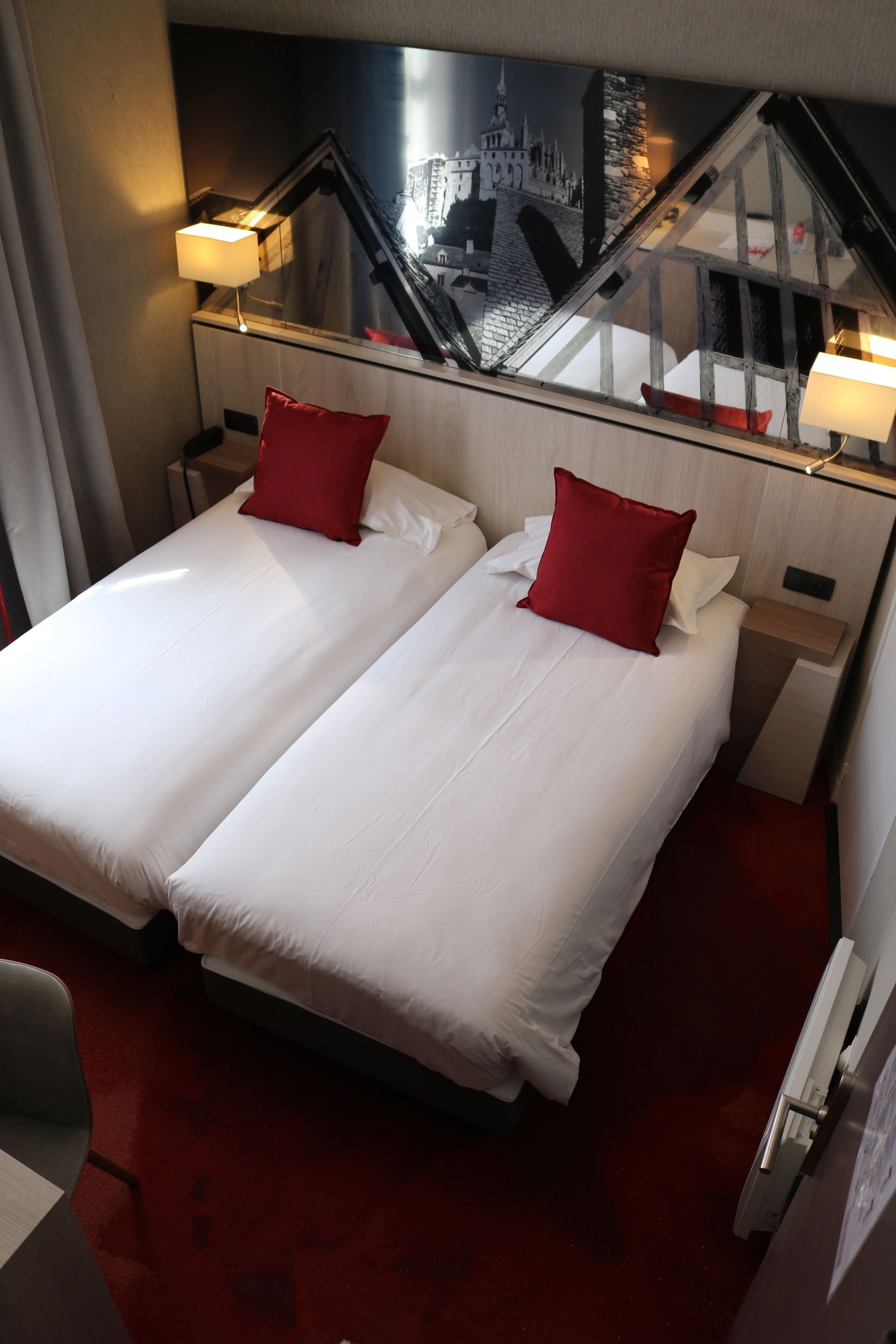 Habitación de hotel con dos camas, almohadas rojas, alfombra roja, vista a la ciudad y una escalera.