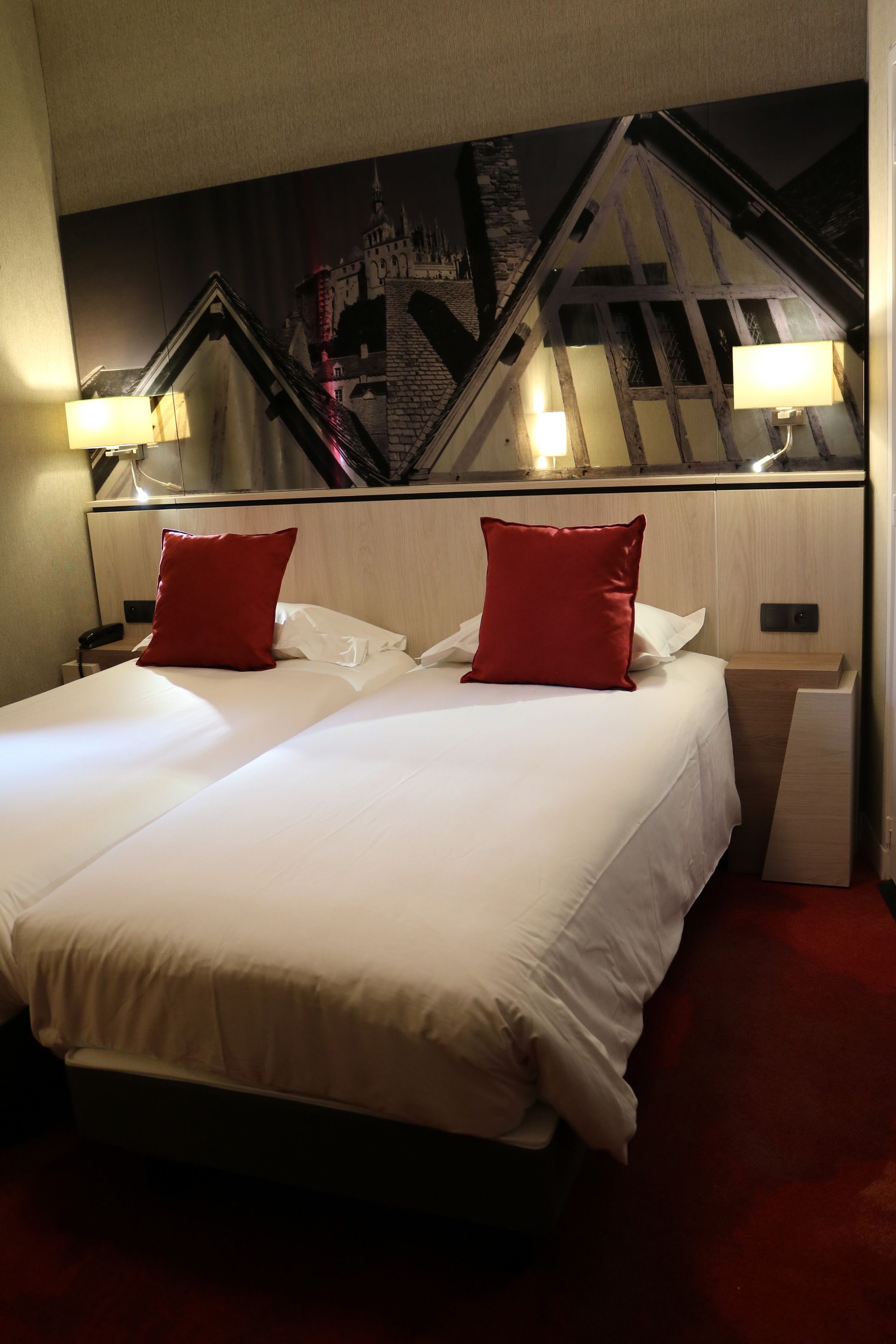 A hotel room with a large bed, two red pillows, a nightstand with a lamp, and a cityscape mural on the wall.