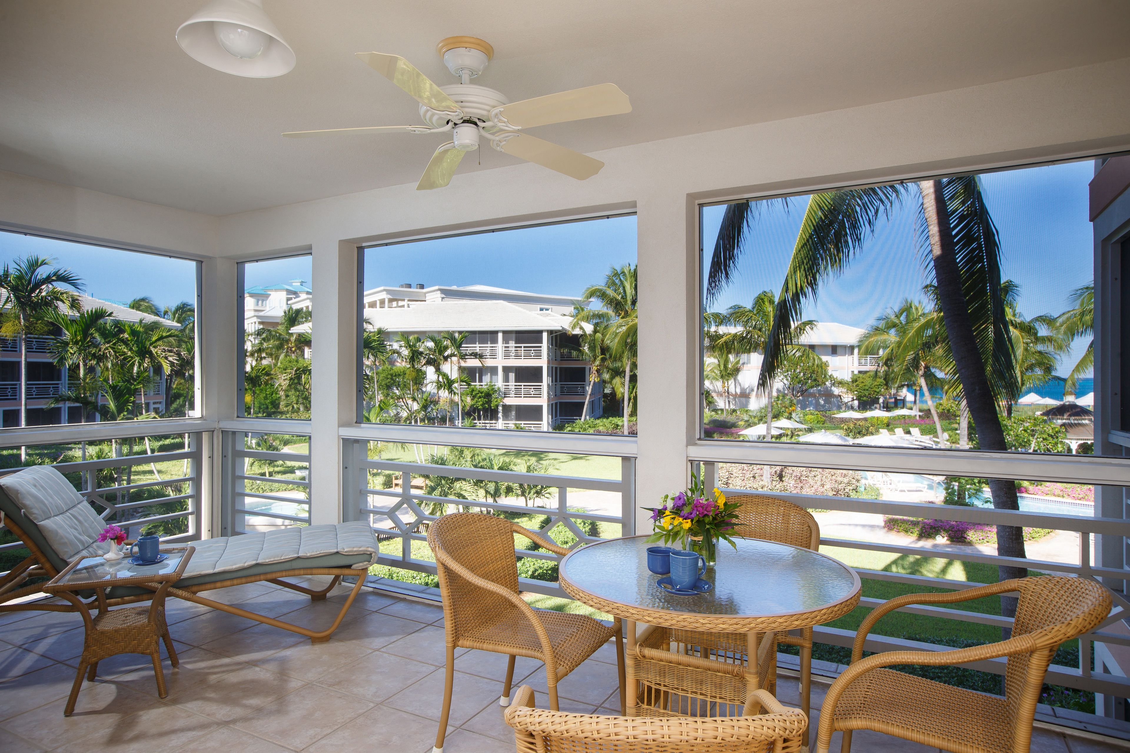 A sunlit room with a ceiling fan, large windows, wicker furniture, and a view of palm trees and buildings.