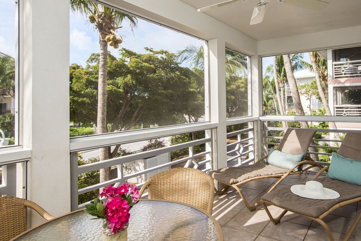 A sunlit balcony with wicker chairs, a table with a vase of flowers, and a view of palm trees and buildings.