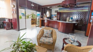 A reception area with a wooden reception desk, two receptionists, wicker furniture, a glass globe with a ship model, and a large window with blinds. - Apartments Maritim Playa - Adults Only (San Bartolomé de Tirajana)