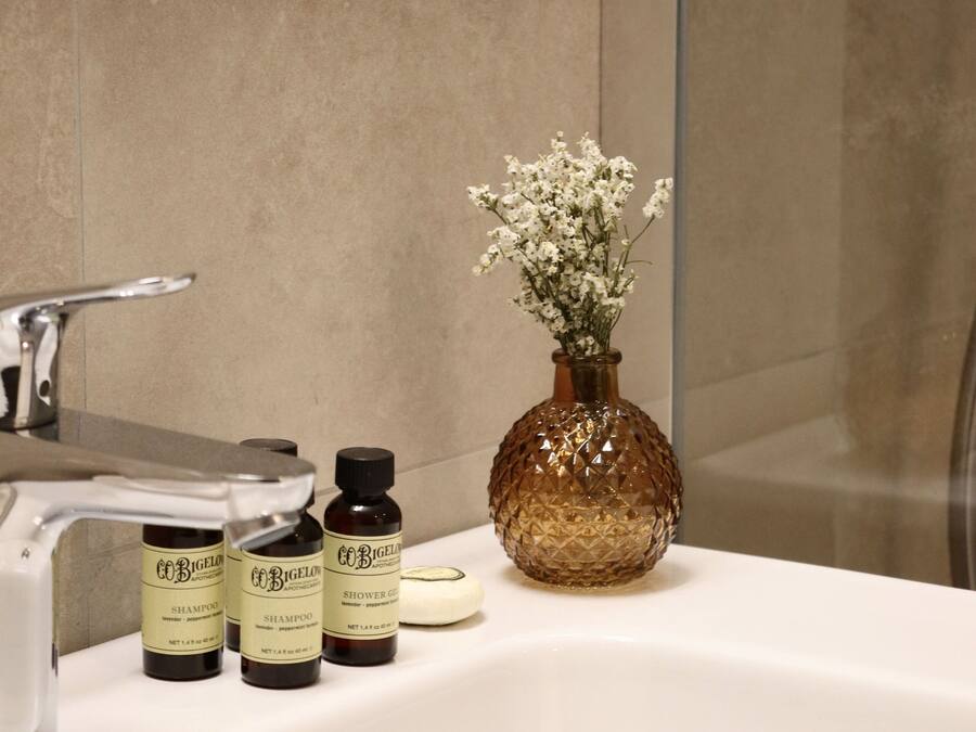 A bathroom with a white sink, three bottles of shampoo, a brown vase with white flowers, and a shower area.