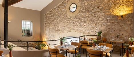 Un restaurante con paredes de piedra, vigas de madera y sillas color naranja. Las mesas están dispuestas con platos, vasos y cubiertos.