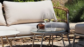 A wicker outdoor seating area with a cushioned sofa, a small round table with two wine glasses and a container of snacks.