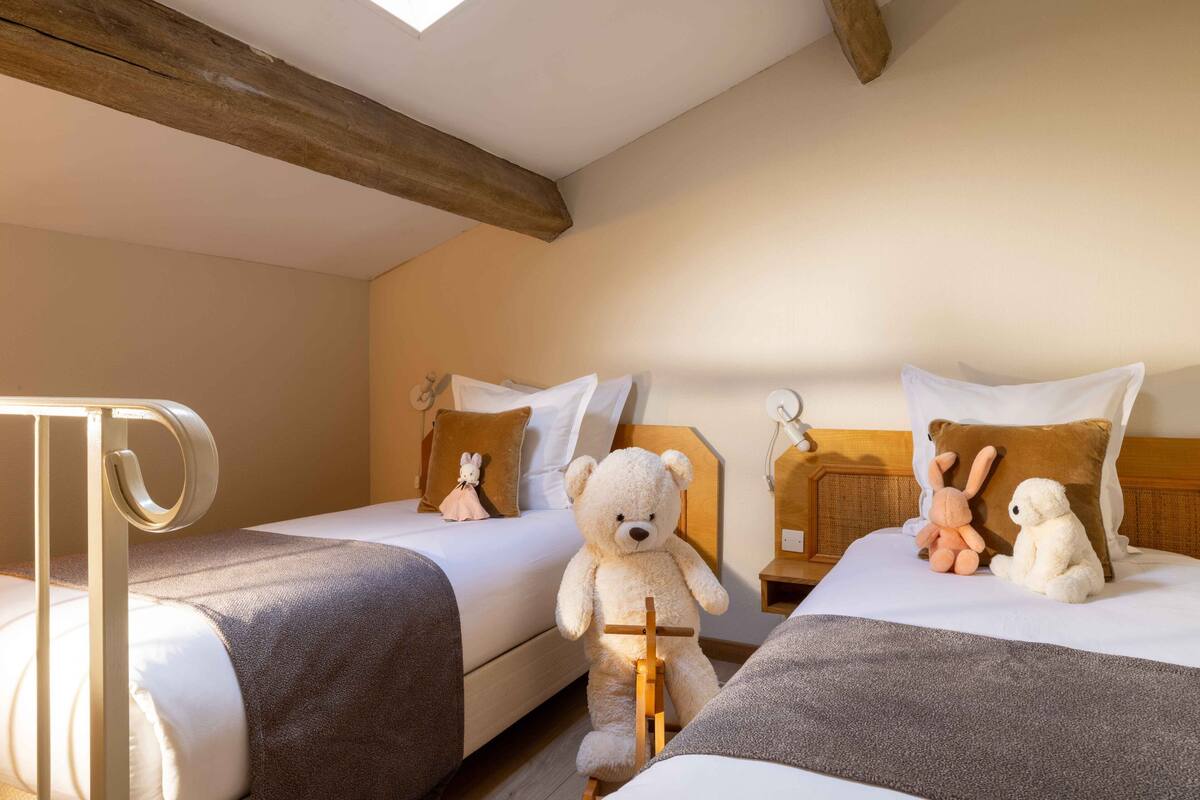 Ein Hotelzimmer mit zwei Betten, jeweils mit Kopfteil, Nachttisch und Lampe. Auf den Betten liegen weiche Kuscheltiere.