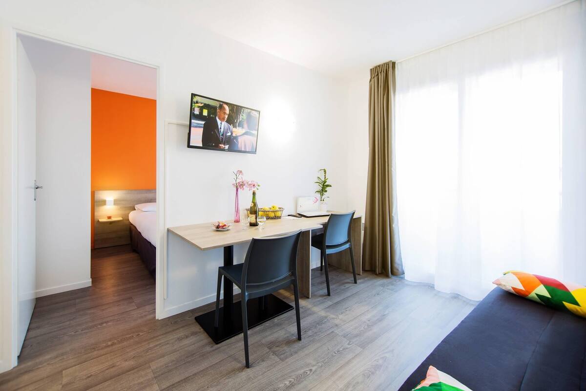 Una habitación moderna con una mesa de comedor, dos sillas, un televisor y una cama con un cojín colorido.