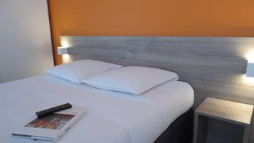 A neatly made bed with white linens and pillows, a remote control, and a magazine on the bed. There is a small bedside table with a lamp on it.