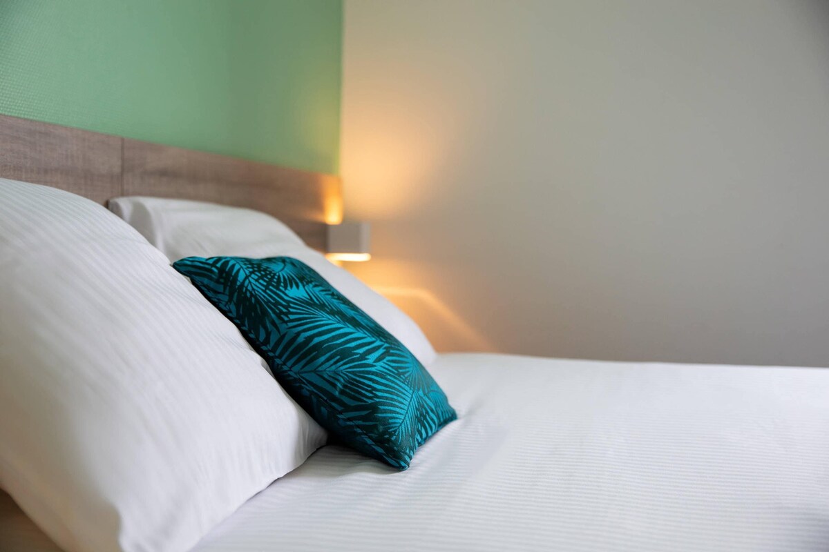 Una cama bien hecha con sábanas blancas y un cojín estampado en tonos aguamarina.