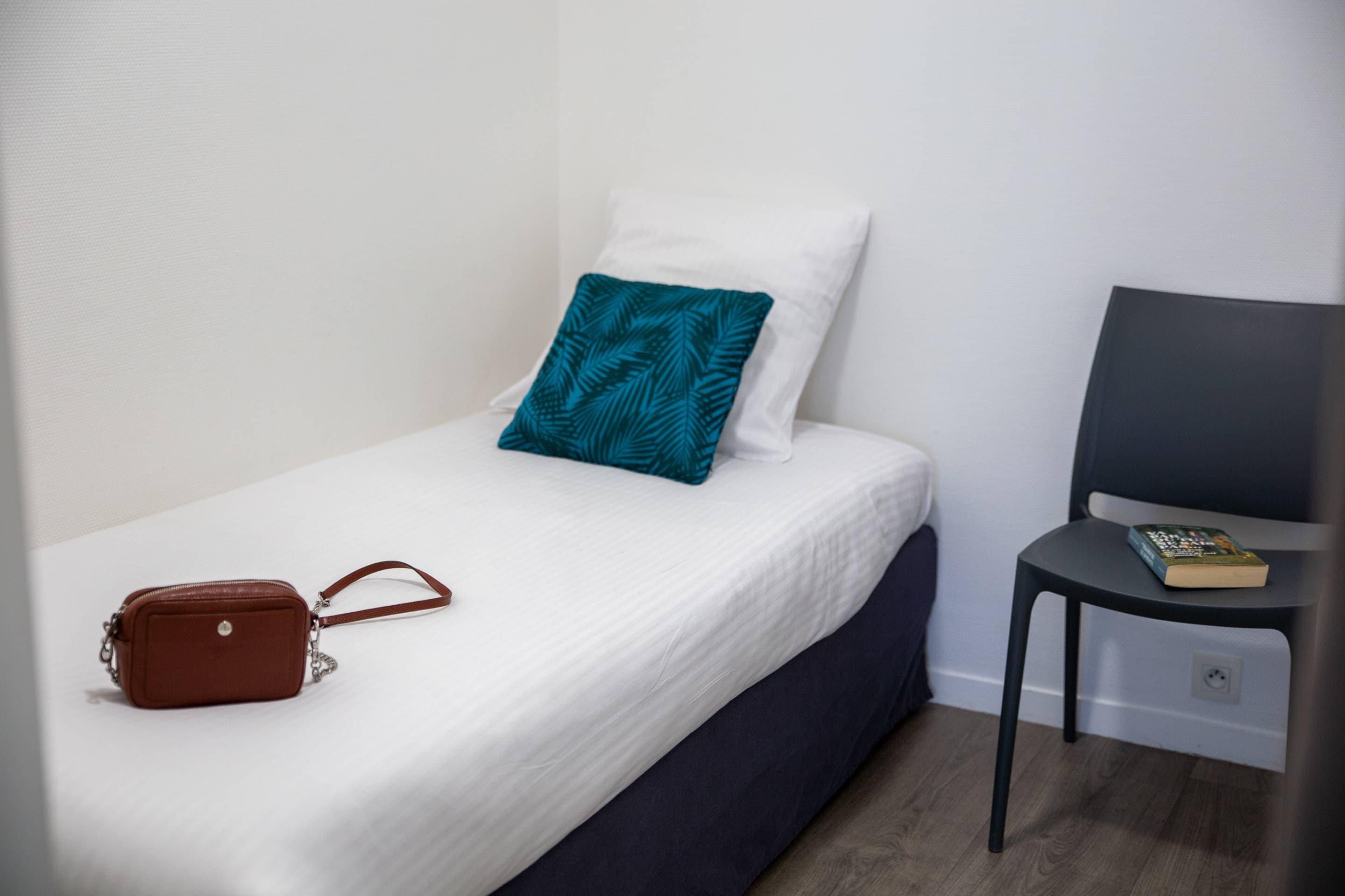Un lit simple avec un matelas blanc et un oreiller turquoise, un sac à main marron posé sur le lit, et une chaise avec un livre.