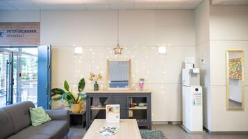 A modern breakfast area with a sign for Petit Déjeuner, a refrigerator, a coffee machine, a small table with brochures, a sofa, and a potted plant.