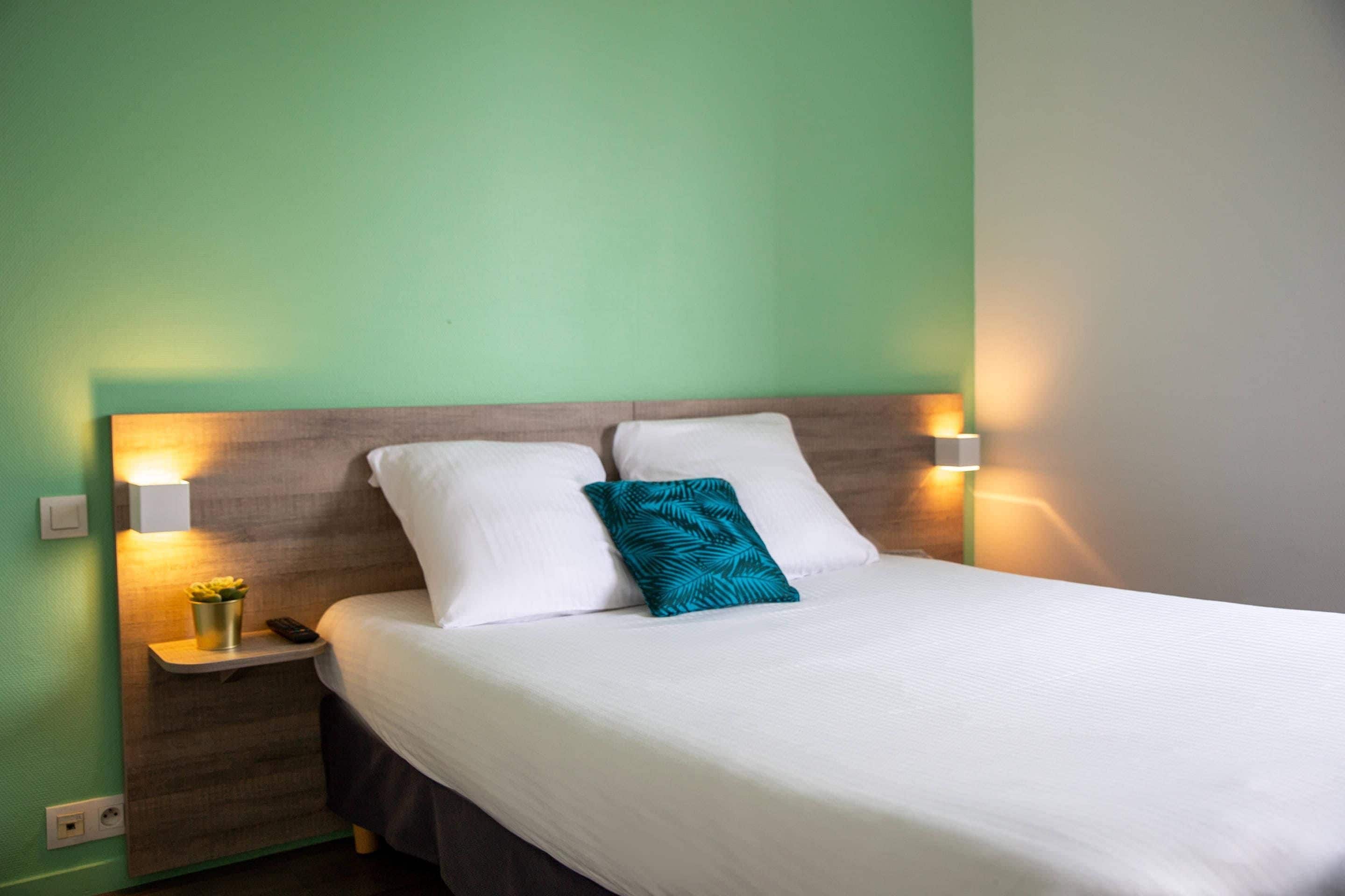 A neatly made bed with white linens and a teal pillow, flanked by two bedside lamps on a nightstand with a small vase of flowers.
