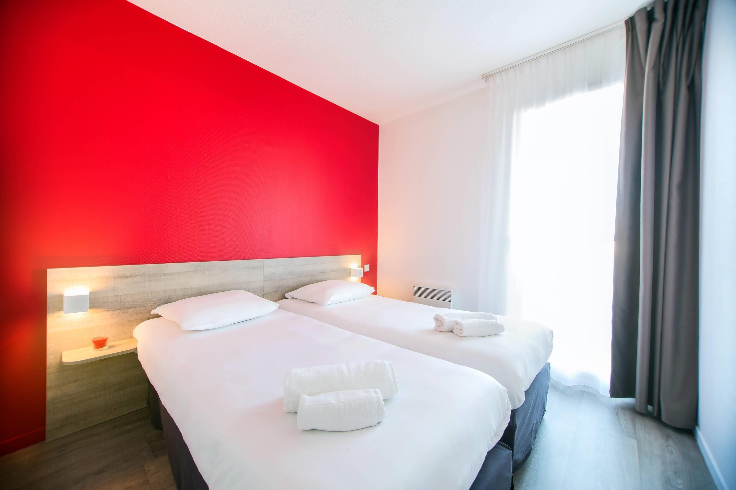 Habitación de hotel con dos camas, una pared decorada en rojo, una ventana con cortinas y una mesita de noche con lámpara.