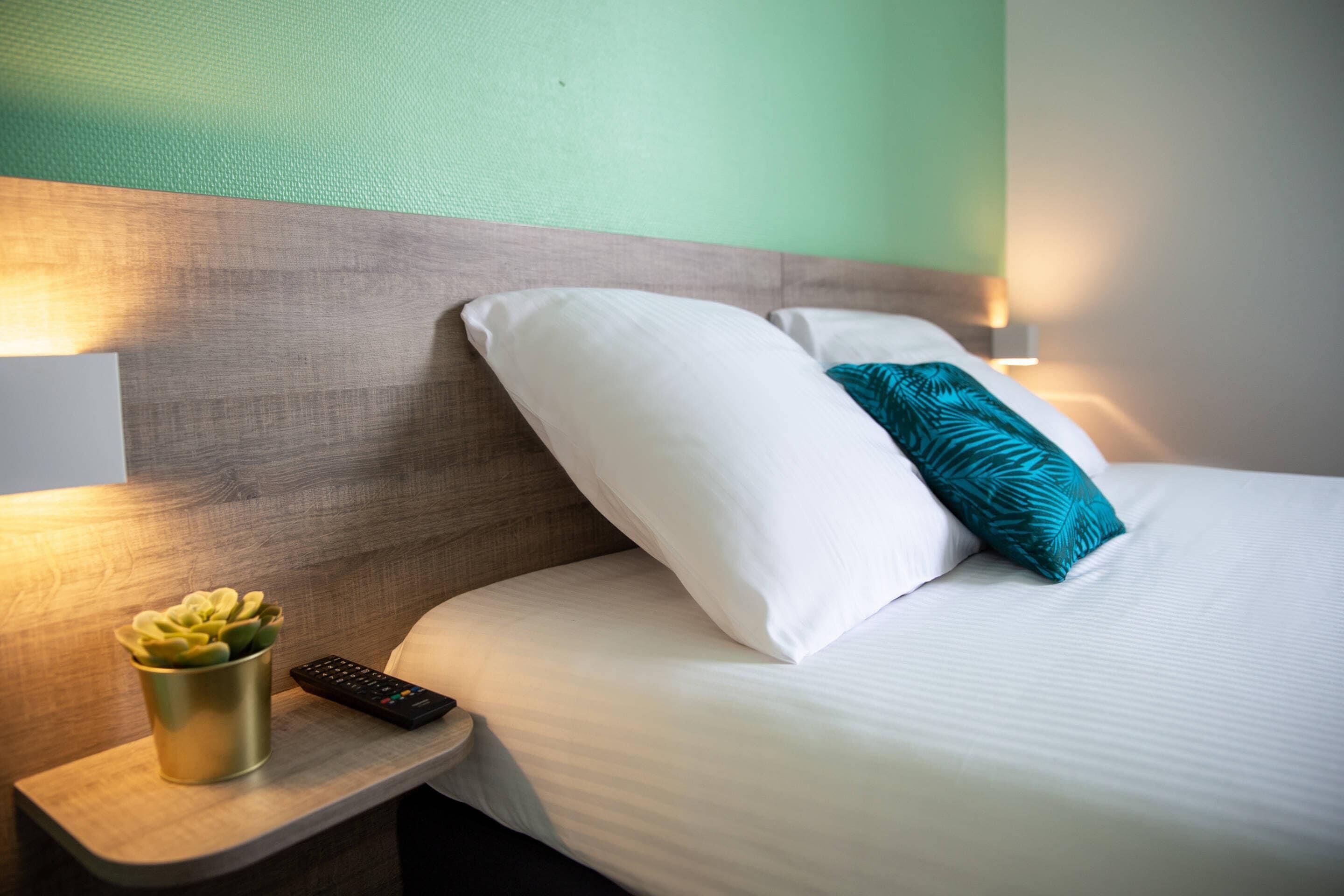 A neatly made bed with white and teal pillows, a remote control, and a small plant on a bedside table.