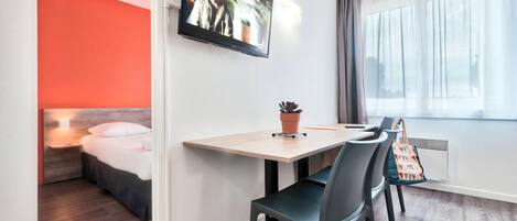 A modern hotel room with a bed, a TV mounted on the wall, a dining table with chairs, and a window with curtains.