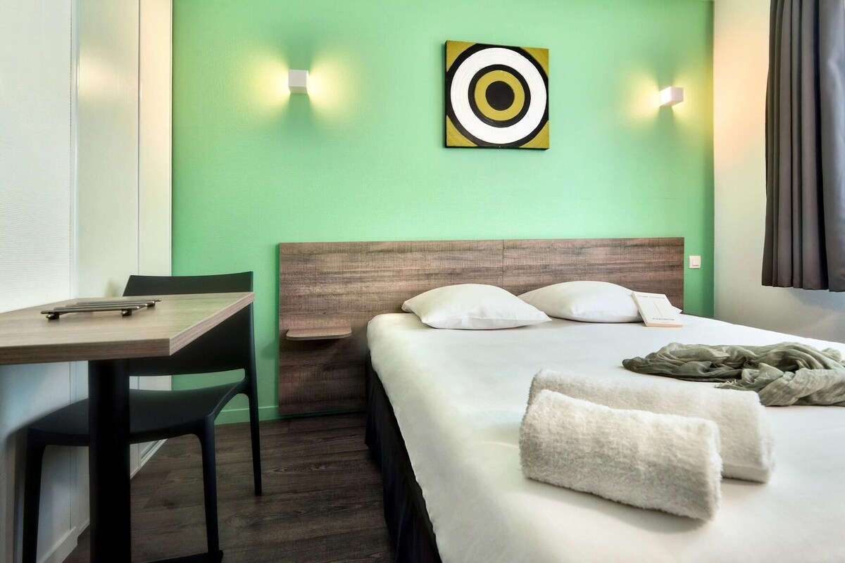 Habitación de hotel con cama, escritorio, silla, cuadro en la pared y ventana con cortinas.