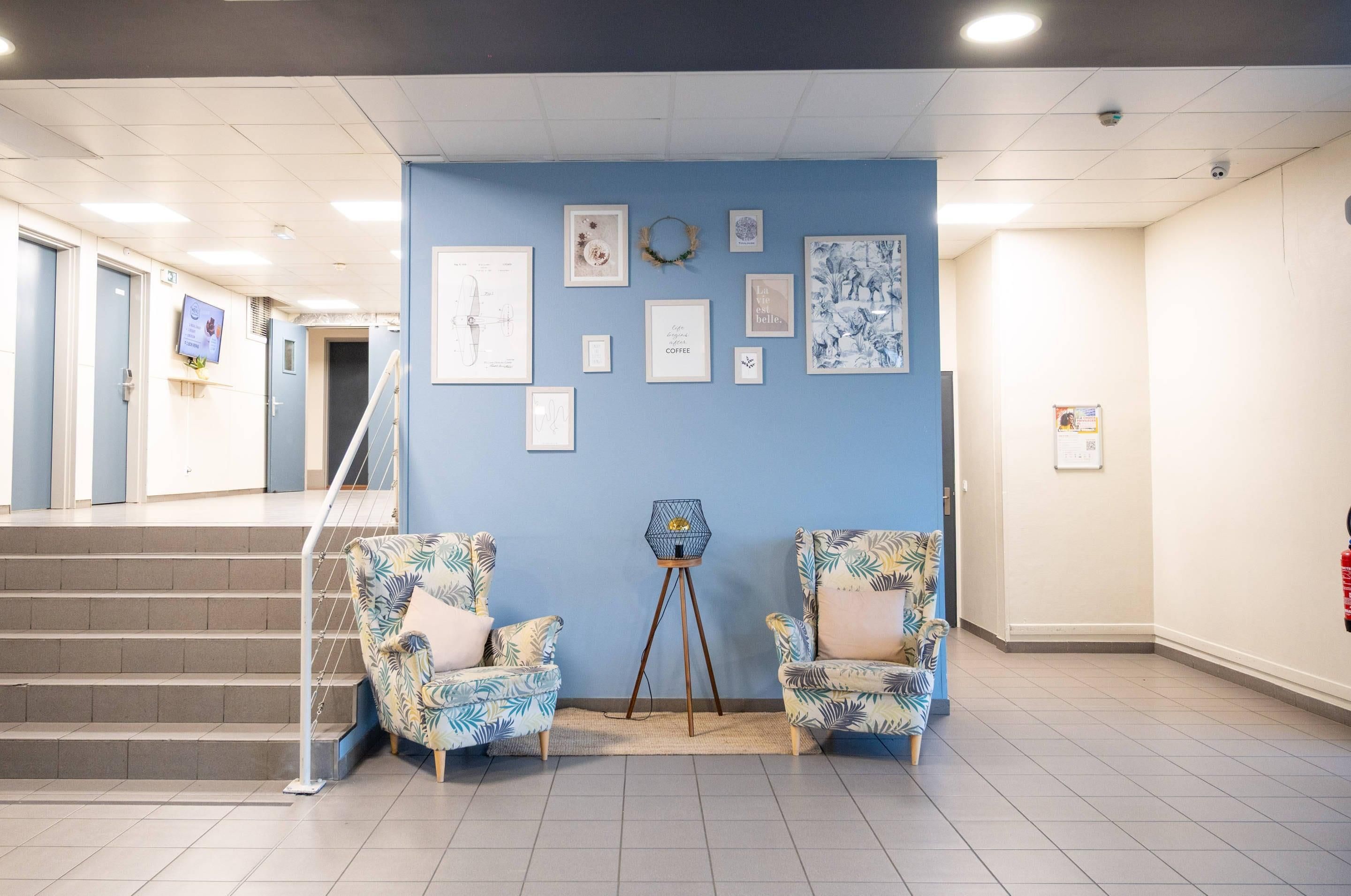 A hallway with a blue wall, framed artwork, two patterned chairs, a small table, and a staircase.