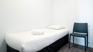 Un lit simple avec une literie blanche et une table de chevet noire à pieds tournés, une chaise sombre et un mur blanc.