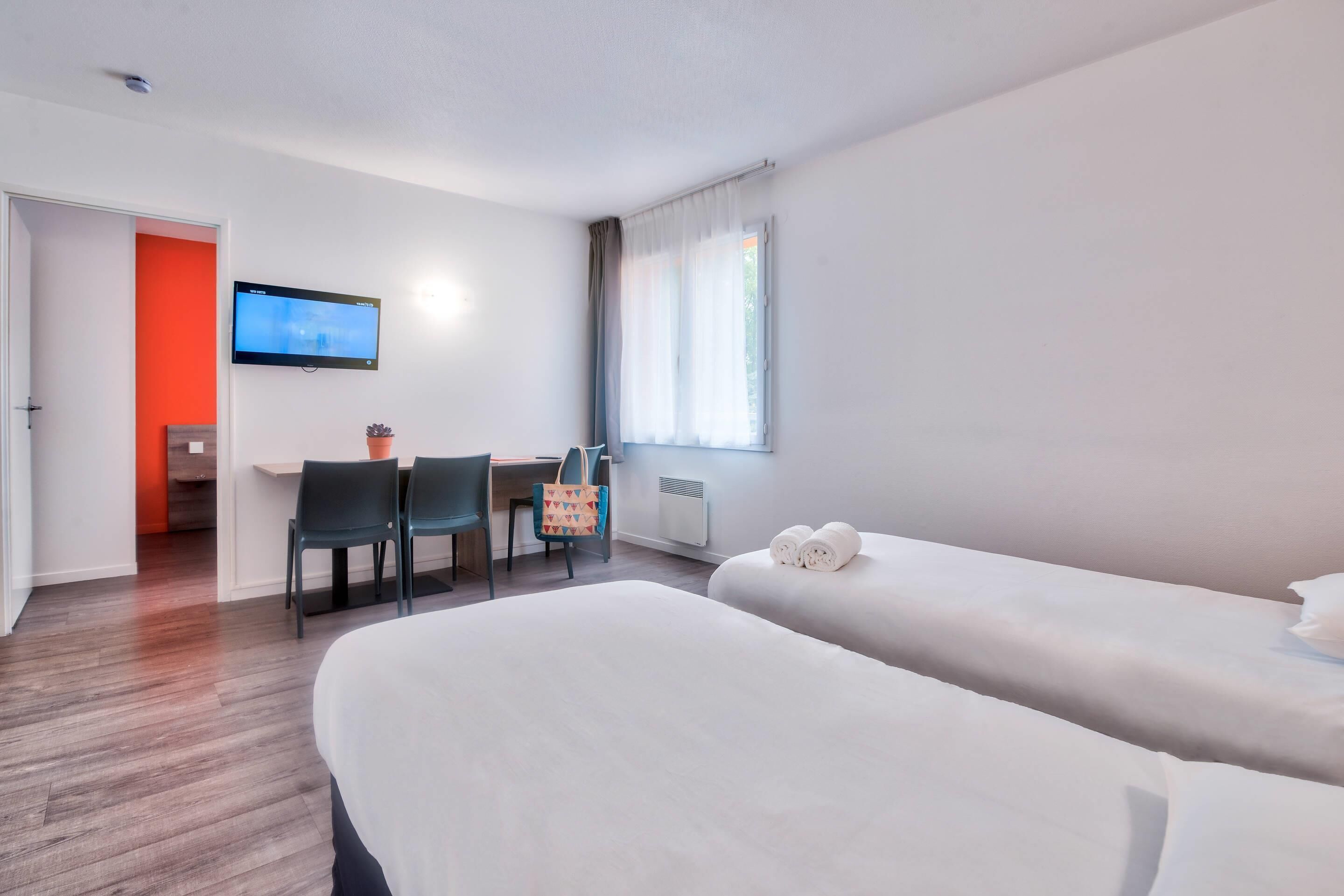 Una habitación de hotel con dos camas, un escritorio con silla, un televisor en la pared y una ventana con cortinas.