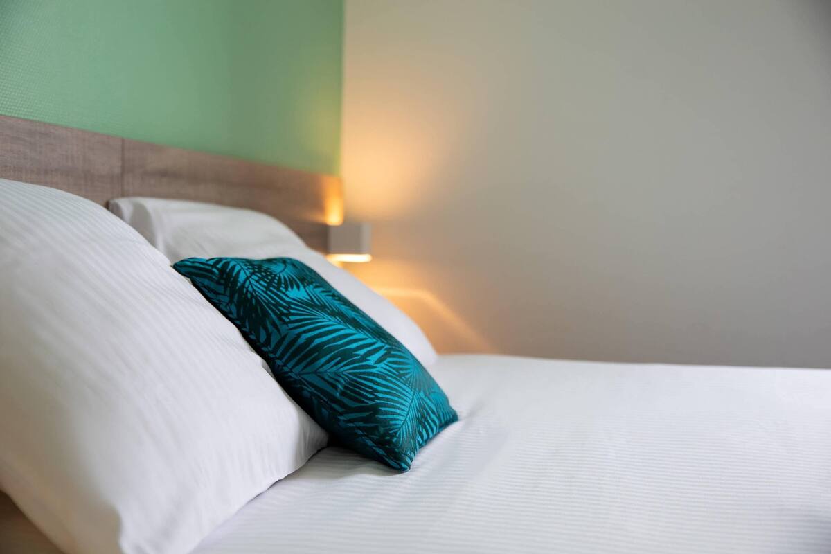 Una cama bien hecha con sábanas blancas y un cojín estampado en tonos aguamarina.