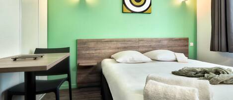 A hotel room with a bed, a desk, and a chair. There is a painting on the wall and a window with curtains.