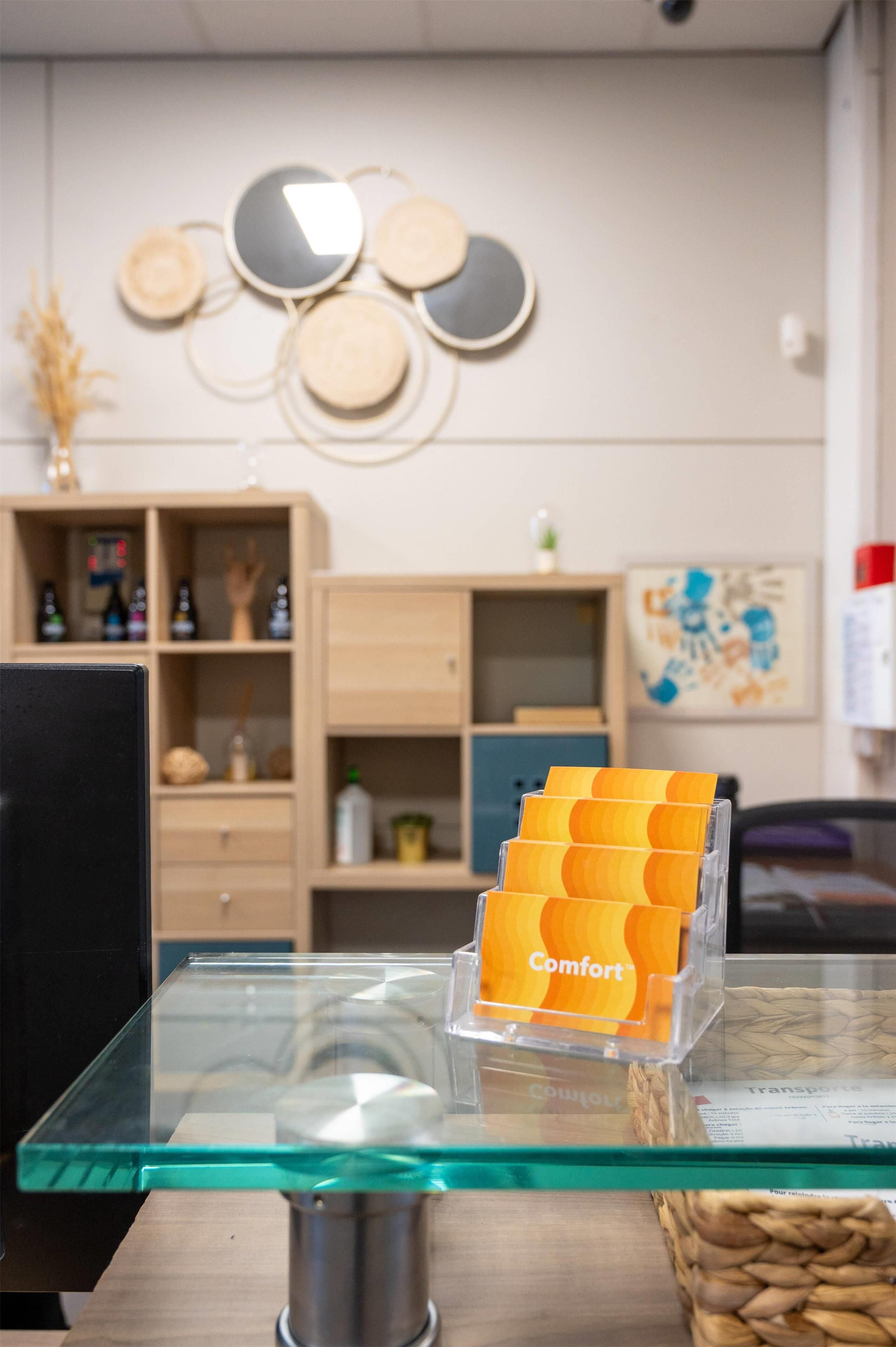 A glass-top table with a clear plastic organizer holding orange cards labeled "Comfort." In the background, there is a wooden cabinet with various items and a wall decoration.