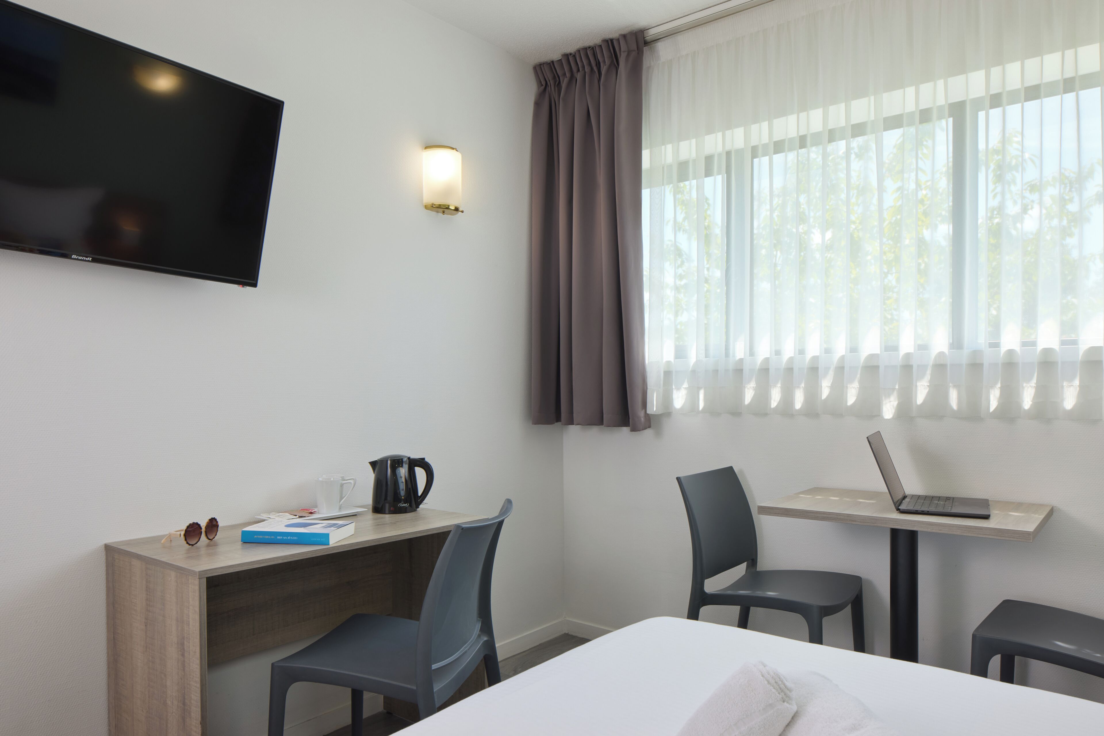 Et hotelværelse med en seng, et skrivebord med en bærbar computer, kaffemaskine og et fjernsyn på væggen.