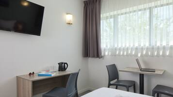 Et hotelværelse med en seng, et skrivebord med en bærbar computer, kaffemaskine og et fjernsyn på væggen.