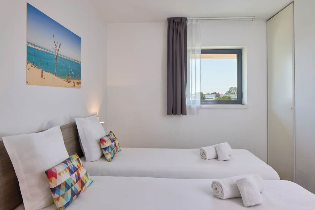 Habitación de hotel con dos camas, una ventana con cortinas, un cuadro de playa y vistas a edificios.