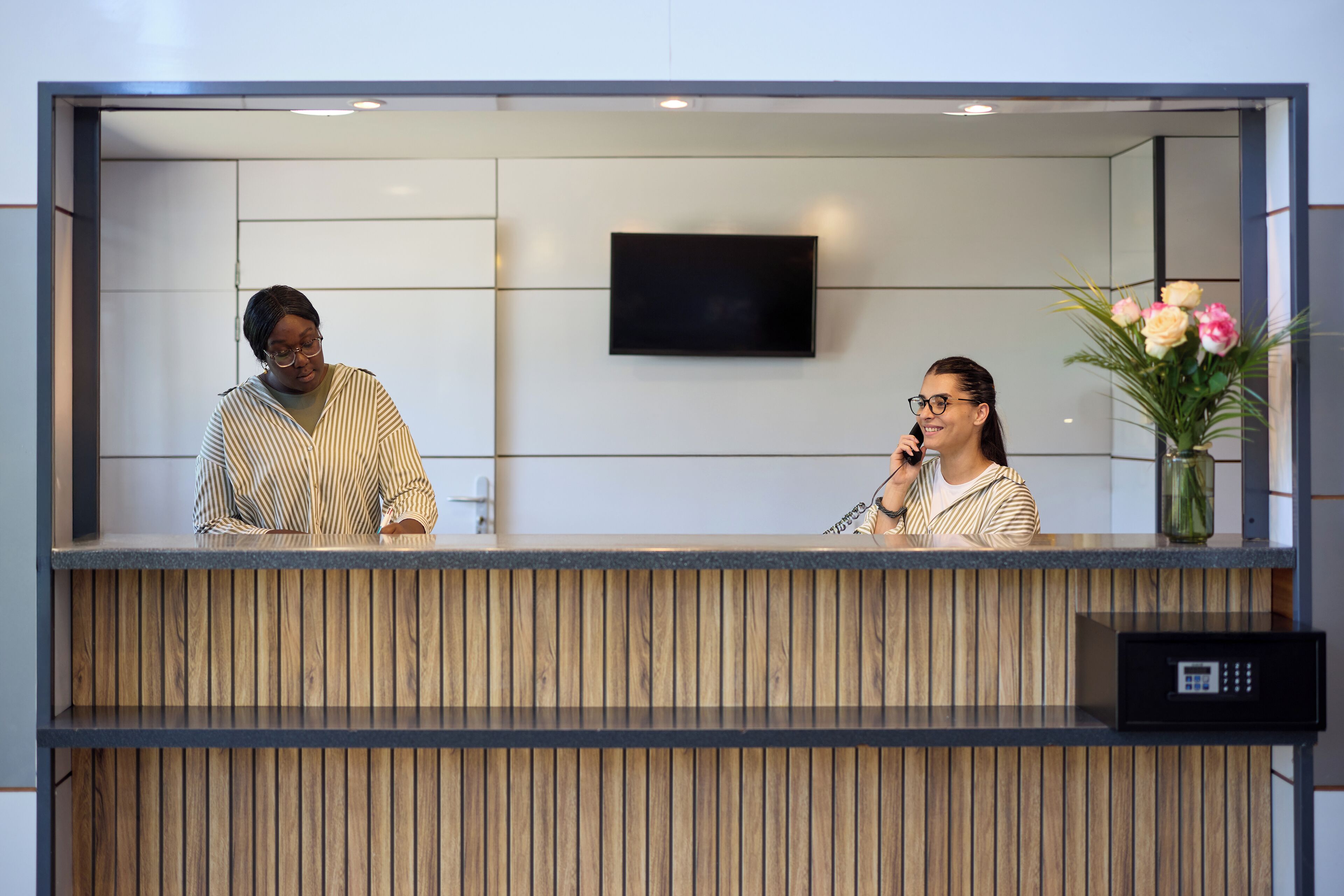 A reception desk with a flat-screen TV mounted above it, a wooden panel design, and a vase with flowers to the right.