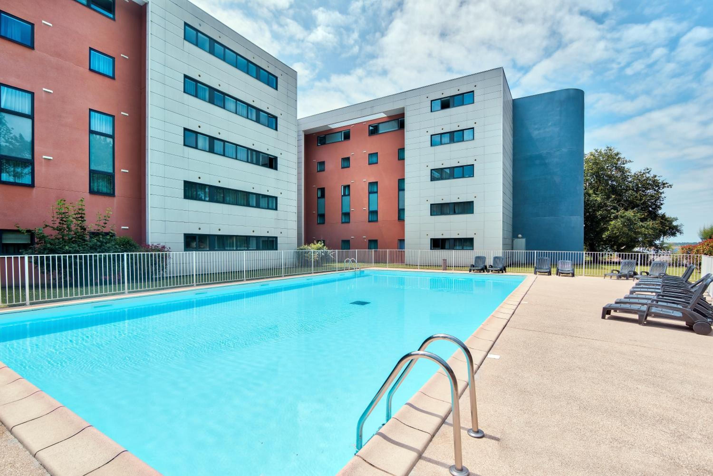 Una piscina con escalera metálica, rodeada por una valla blanca, y contigua a un edificio moderno con múltiples ventanas y balcones.