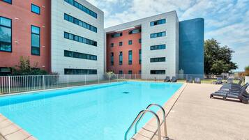 Una piscina con escalera metálica, rodeada por una valla blanca, y contigua a un edificio moderno con múltiples ventanas y balcones.