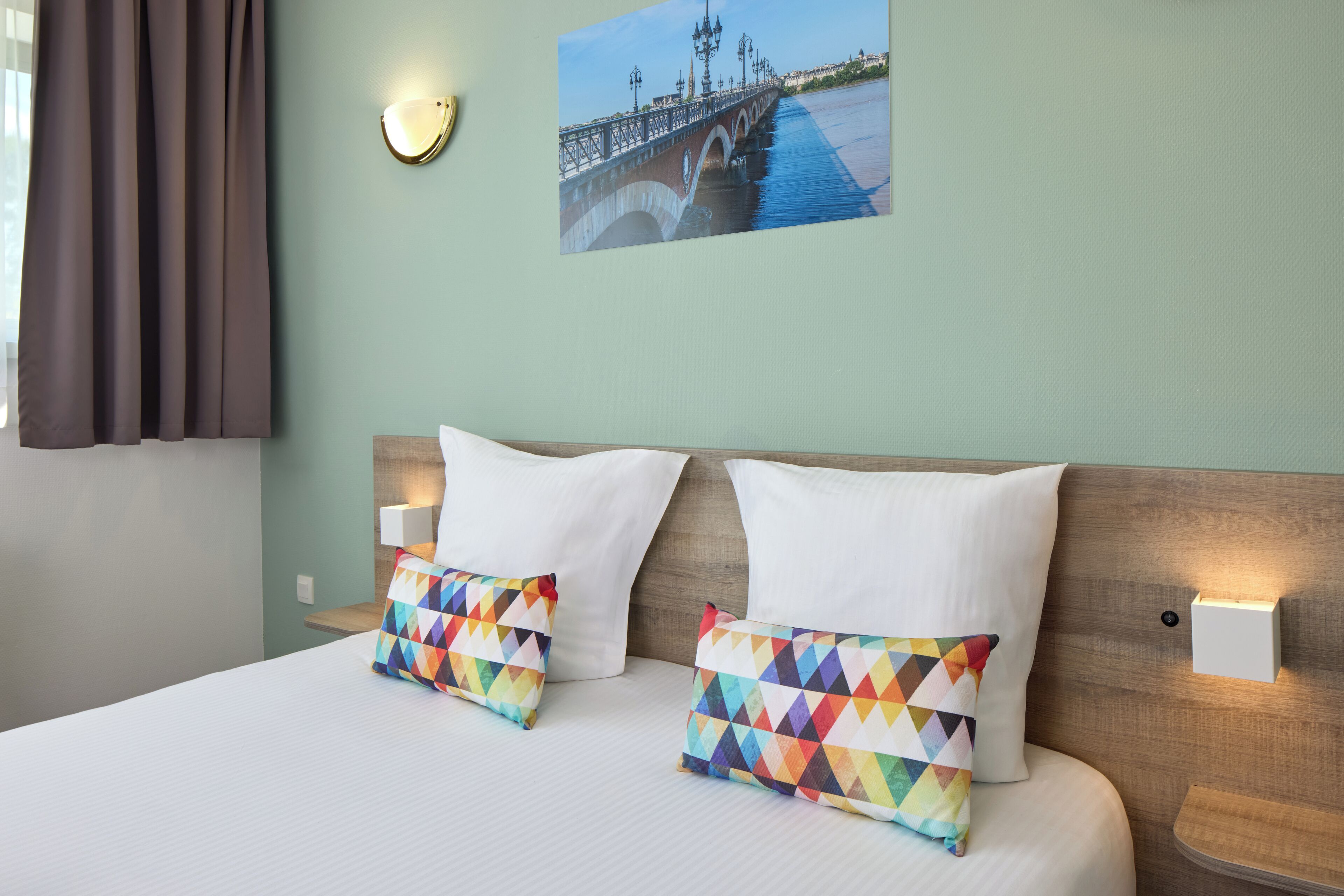 Une chambre d’hôtel avec un lit, deux oreillers blancs, un oreiller décoratif aux motifs géométriques et colorés, une table de chevet avec une lampe, un luminaire fixé au mur et un tableau représentant un pont.