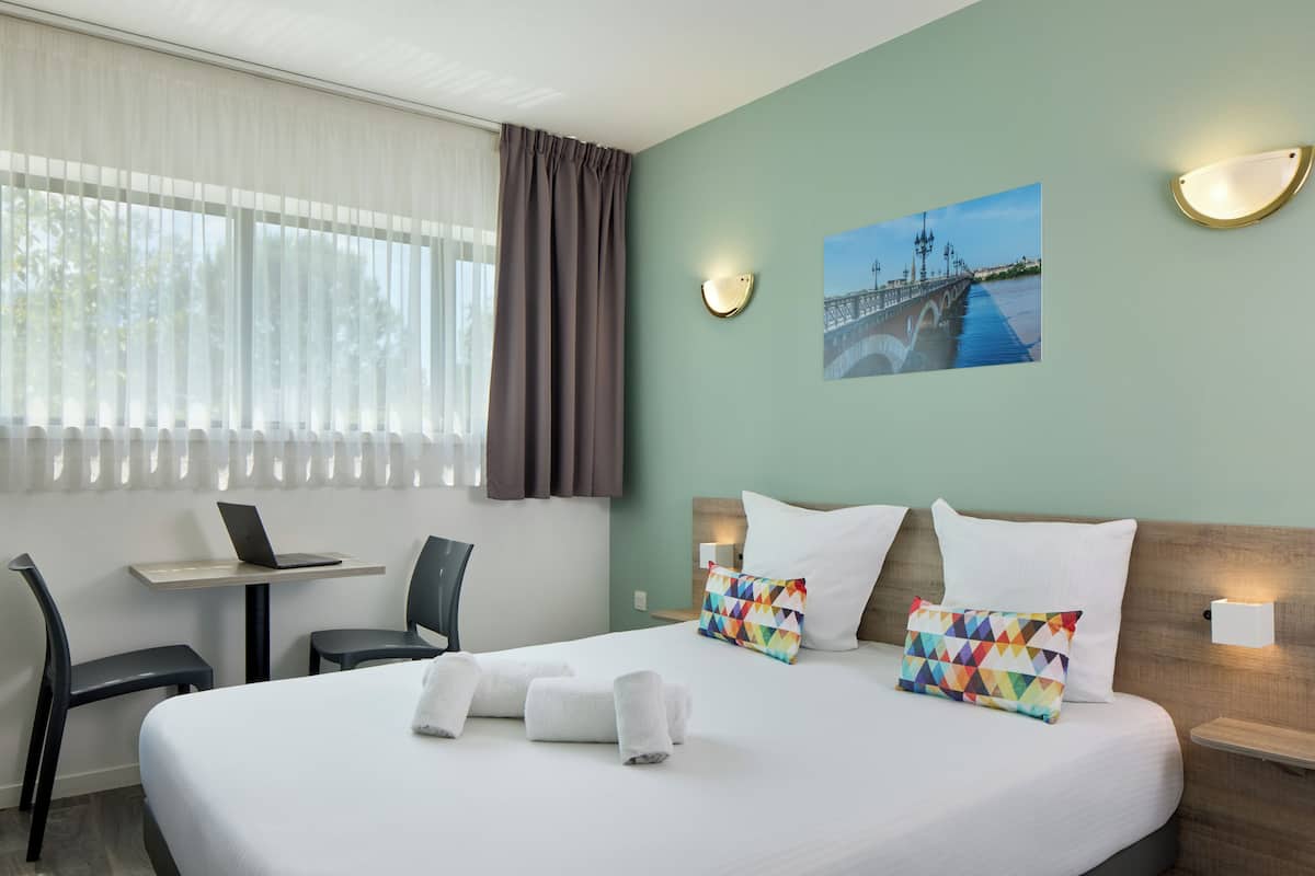 Habitación de hotel con cama, escritorio, computadora portátil, silla y un cuadro en la pared.