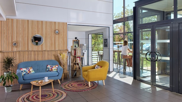 A modern interior with a blue sofa, yellow armchair, and a wooden panel wall. - Zenitude Hôtel - Résidences Bordeaux Aéroport (Merignac)