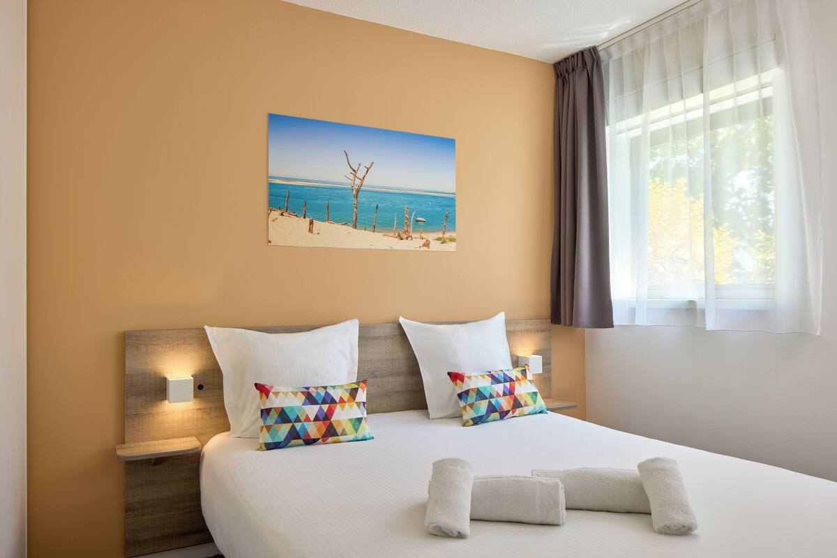 Una habitación de hotel con una cama grande, dos almohadas, una mesita de noche con una lámpara, un cuadro de una playa y una ventana con cortinas.