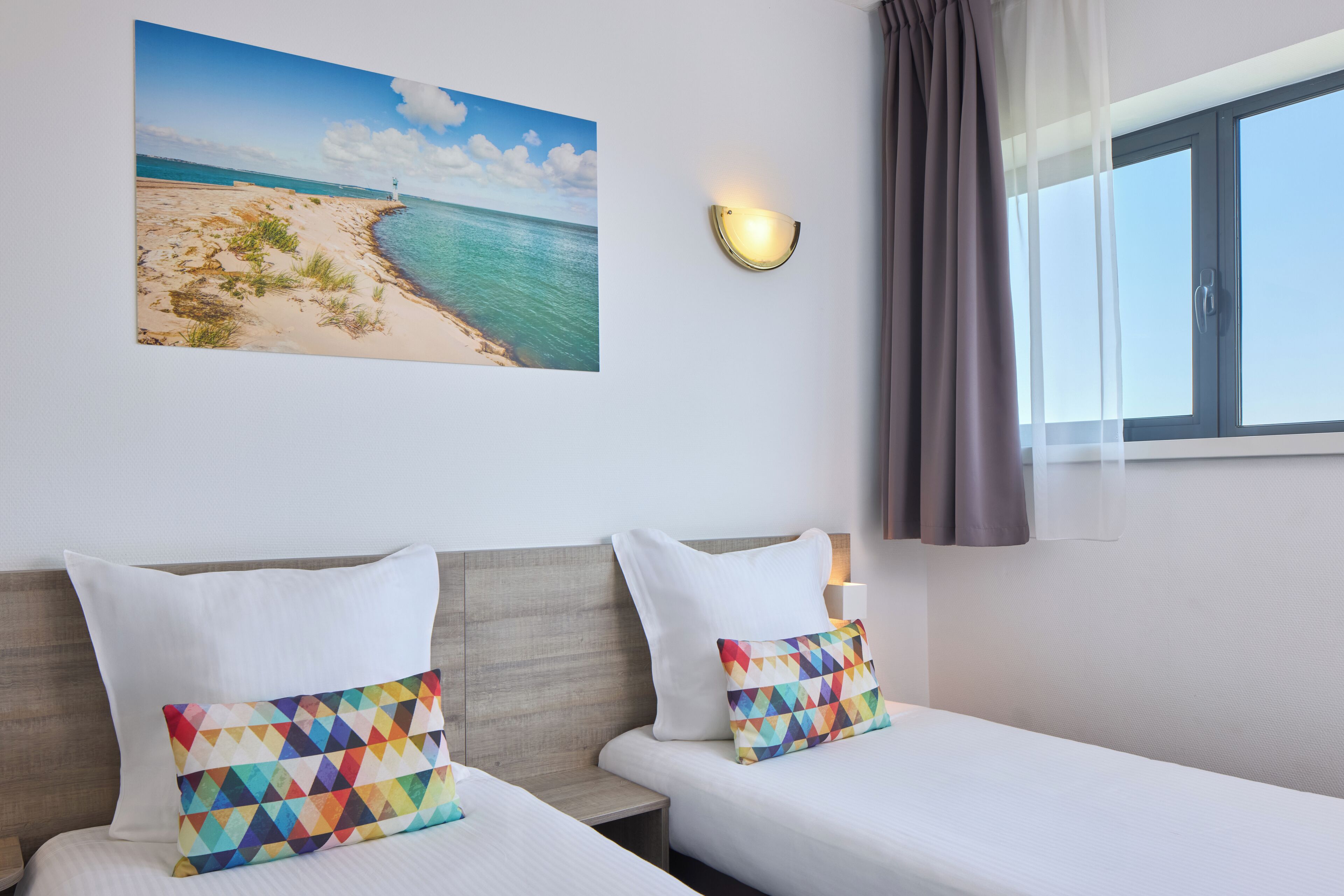 Une chambre d’hôtel avec deux lits, un tableau représentant une plage, un lampadaire fixé au mur et une fenêtre avec des rideaux.